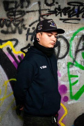 Portrait of refugee Mateo wearing a cap standing against a graffiti wall