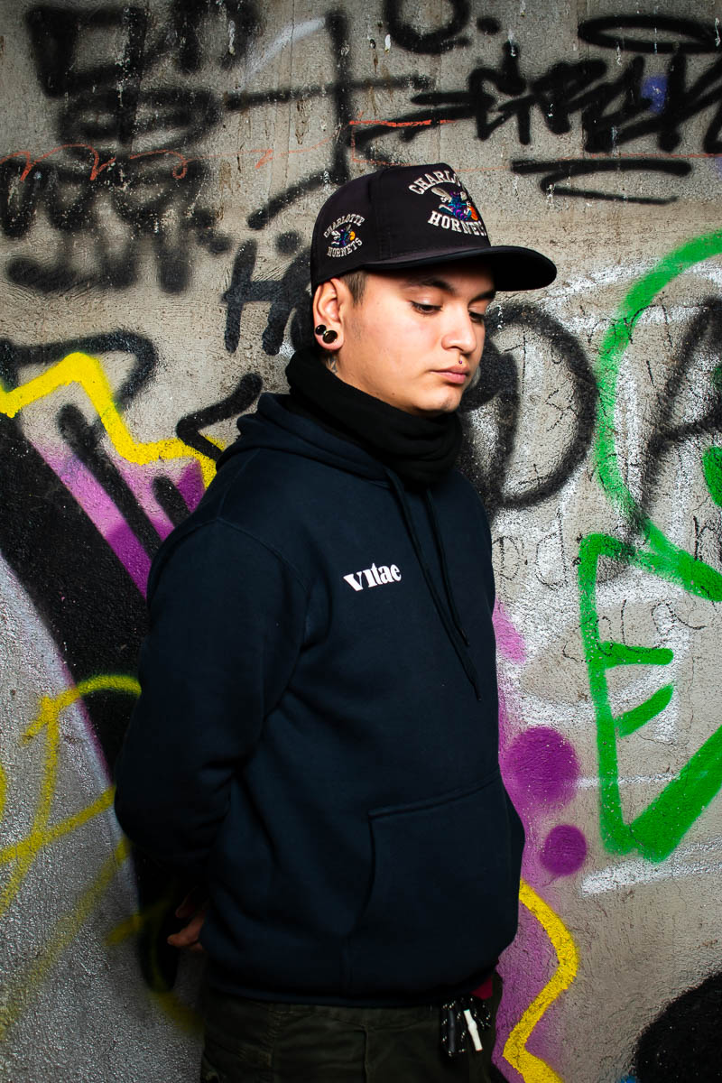 Portrait of refugee Mateo wearing a cap standing against a graffiti wall