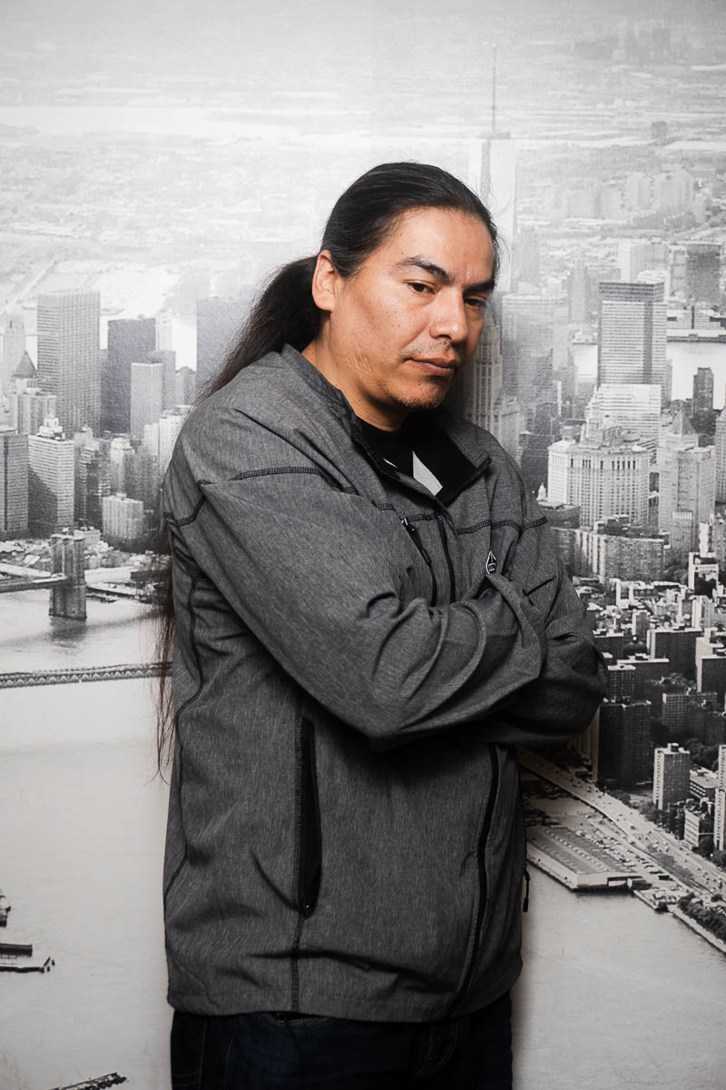 Portrait of Erik, has long hair and is wearing a dark jacket clasping his arms together not facing directly to the camera