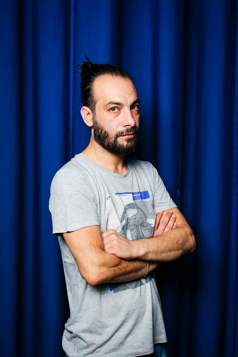 Portrait of Ghazwan looking intently in the camera, hands clasped on stomach and is wearing a grey shirt.