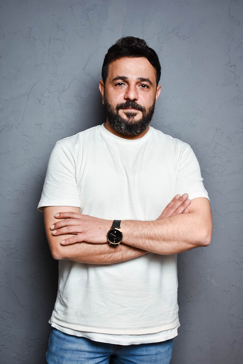 Portrait of Emad, facing the camera clasping his arms together, wearing a watch and a white shirt.