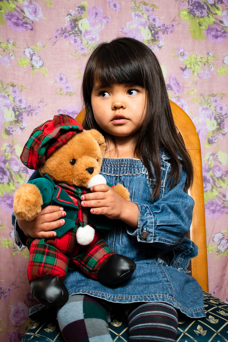 Portrait of Mogjan wearing a denim baby dress holding a stuff toy.