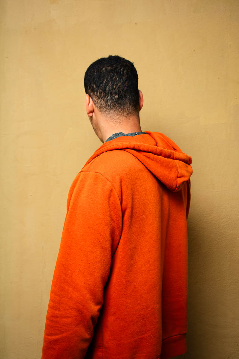 Portrait of Omar, facing at the back, not showing his face and is wearing an orange jacket with a hood.