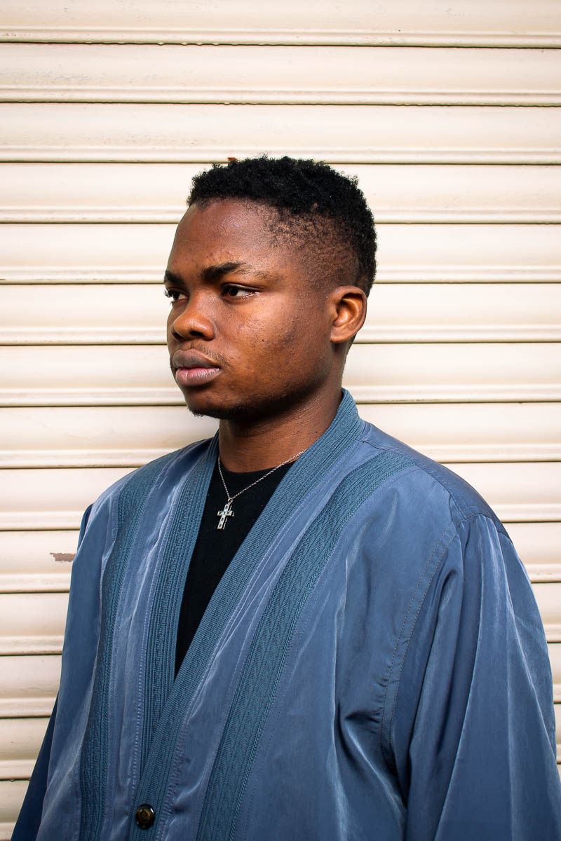 Portrait of Damilare, facing sideward from the camera wearing a blue robe and a black shirt underneath with a cross-styled necklace.