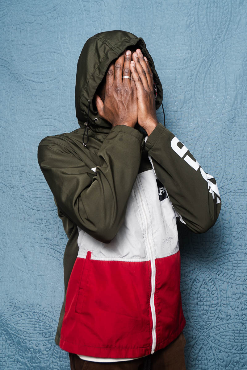 Portrait of Moustapha, covered his face using his hands and is wearing a dark green and red jacket.