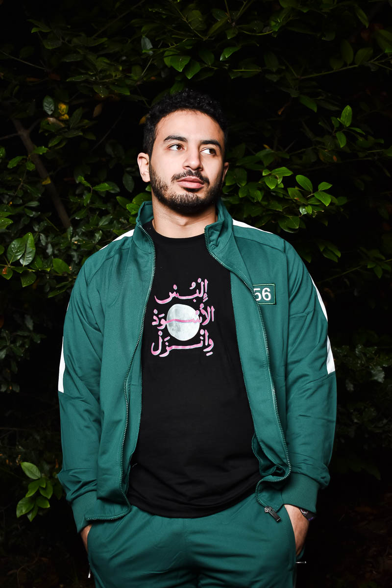 Portrait of Musa, facing sideways, and is wearing a dark green jacket and black shirt underneath.