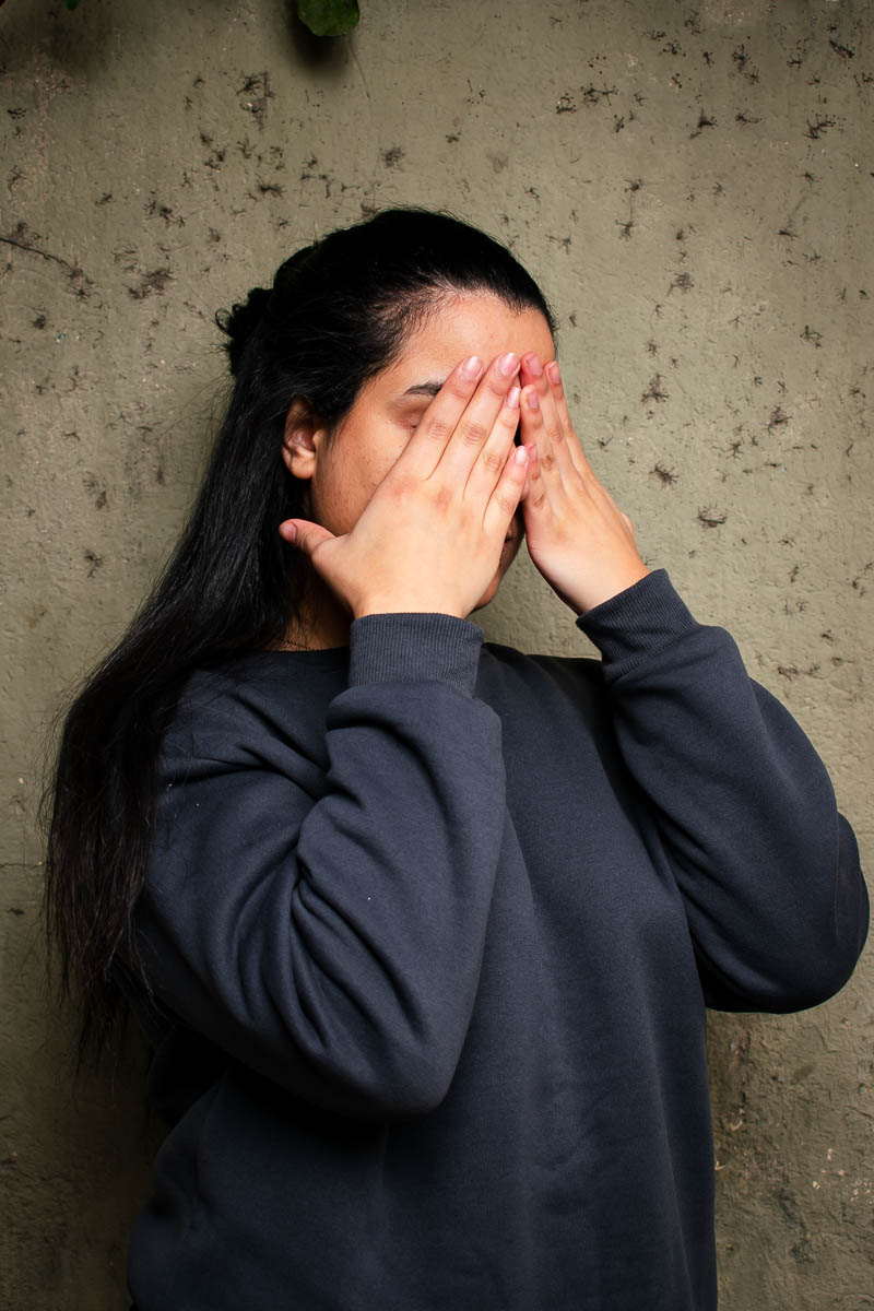 Portrait of Salma, covering her face with her hands and is wearing a black jacket.