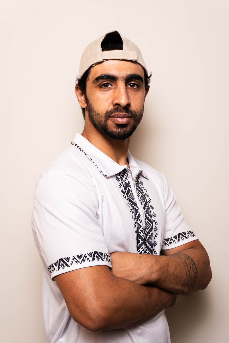 Portrait of Ahmad, looking directly at the camera, arms crossed and is wearing a white polo and a brown cap.