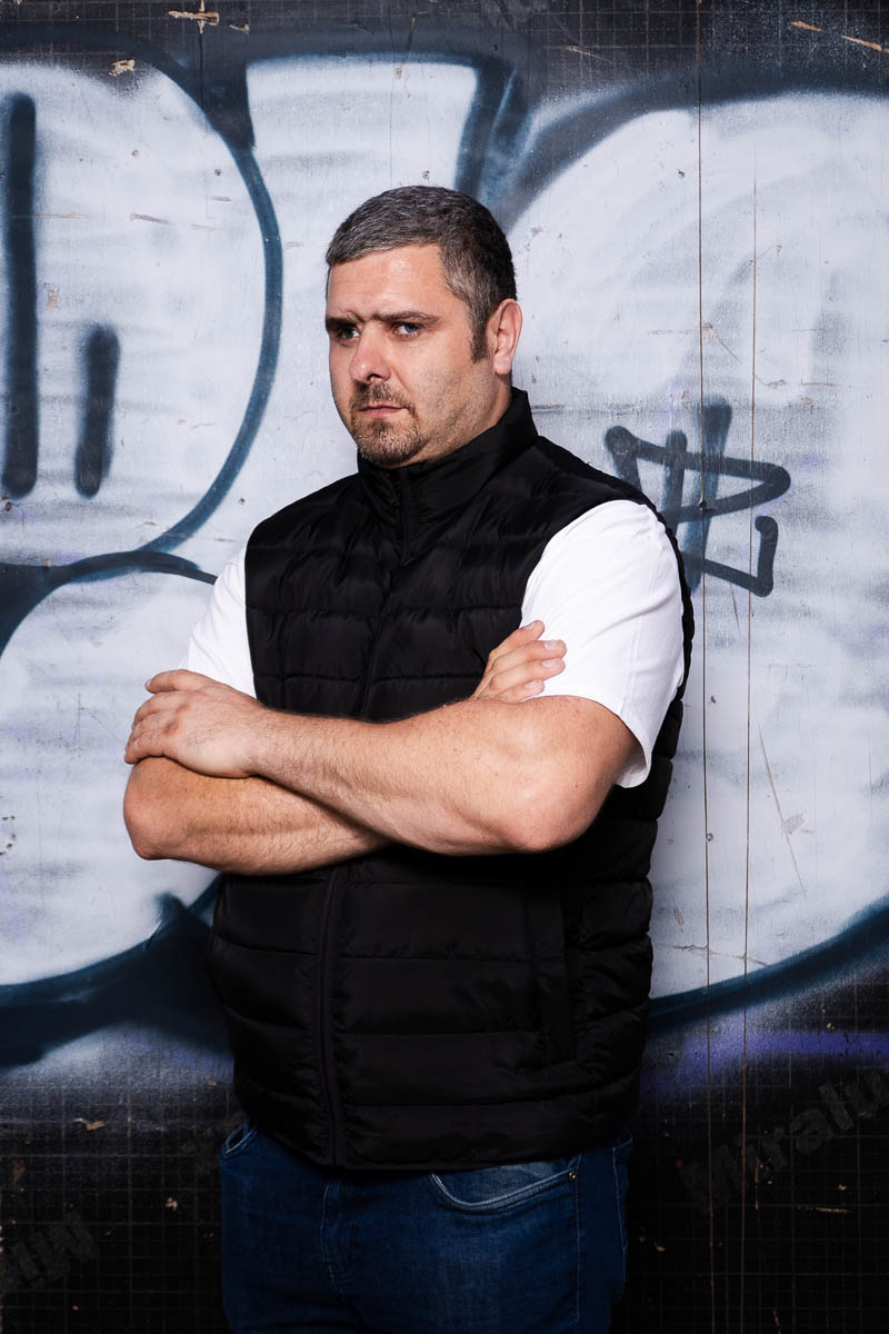 Portrait of Viktor, arms clasped, not looking directly to the camera and is wearing a black suit with white shirt underneath.