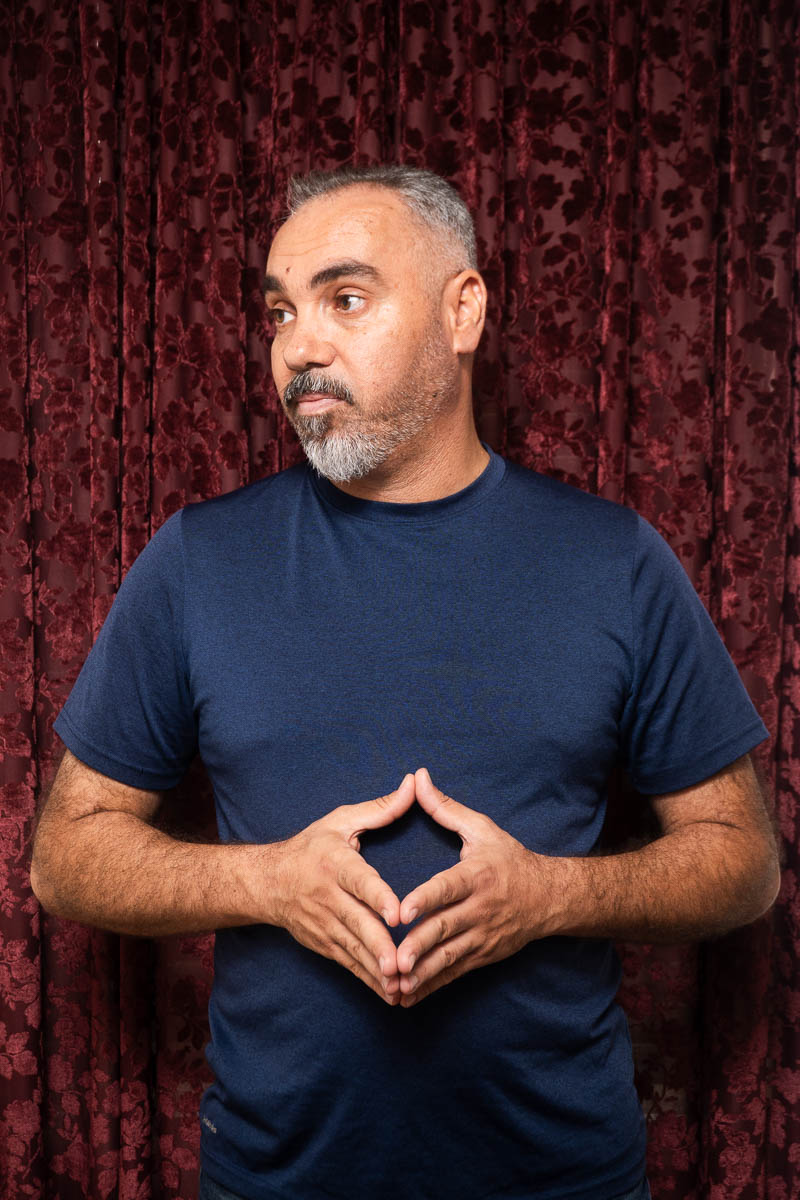 Portrait of Muhanad, hands are clasped on stomach and is wearing a dark blue shirt.