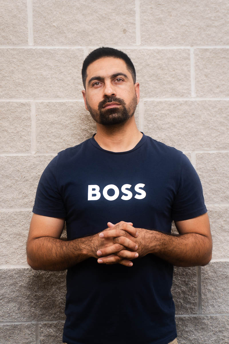 Portrait of Wahid, facing neutral in the camera, hands clasped together in chest and is wearing a dark shirt labeled "BOSS."