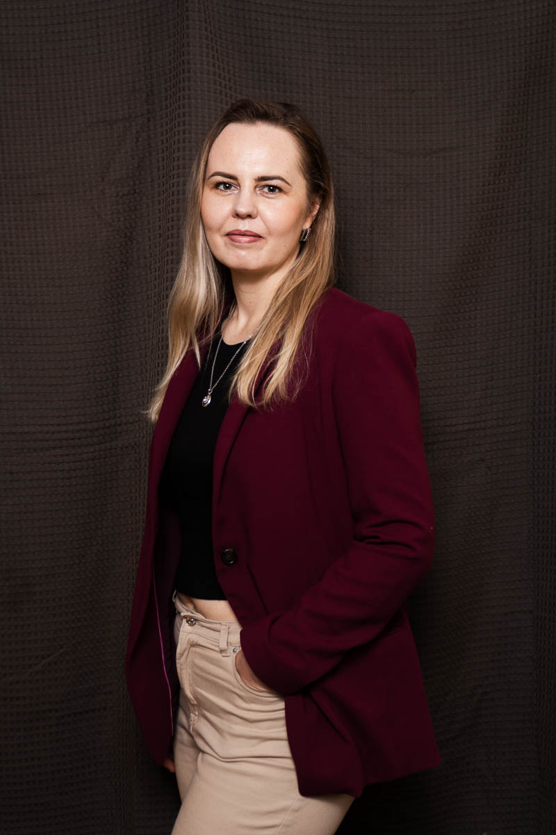 Portrait of Nastya, wearing a maroon suit, hands on purse and is looking directly in the camera.