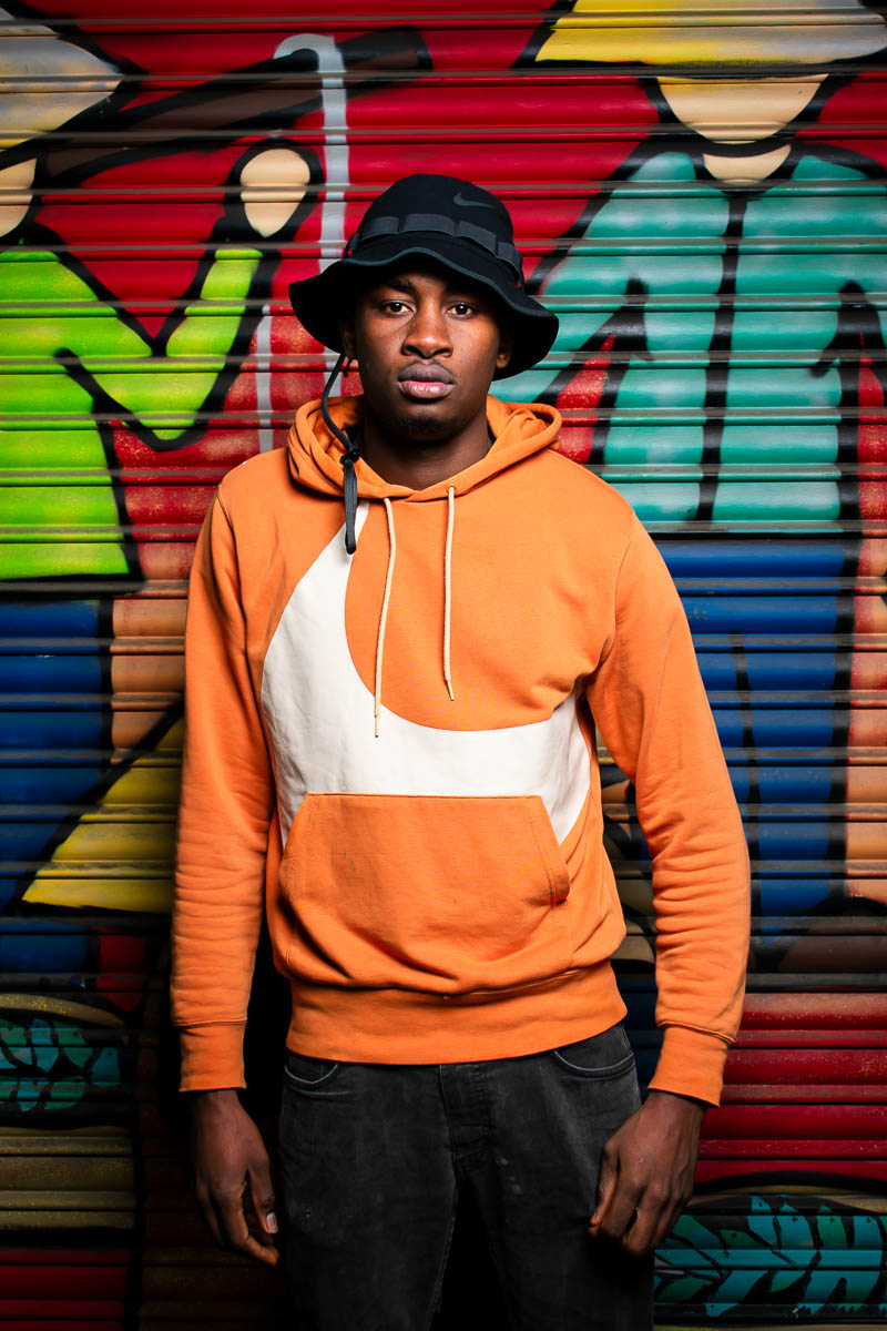 Portrait of Tapha, faving the camera and is wearing an orange jacket with black cap.